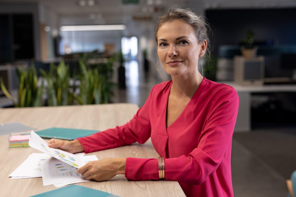 Portrait of smiling caucasian female advisor sitting with document at table in modern workplace